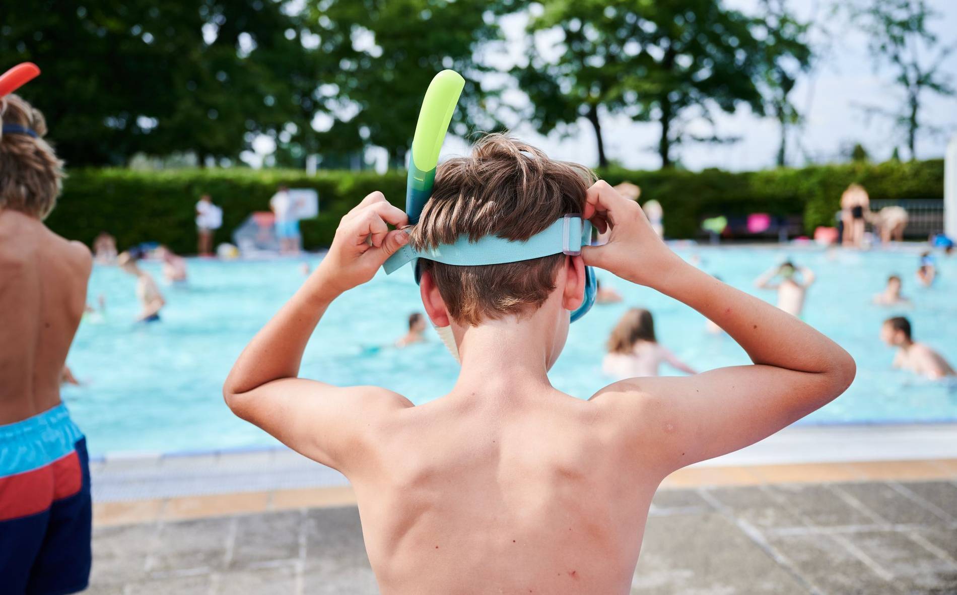 Junge von hinten im Schwimmbad zieht sich seine Taucherbrille an und geht Richtung Schwimmbecken