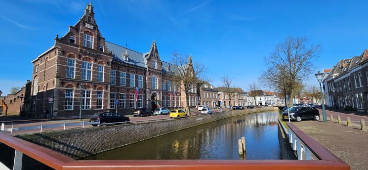 Blick von einer Br&uuml;cke auf einen Kanal in einer niederl&auml;ndischen Kleinstadt, links ein gro&szlig;es Backsteingeb&auml;ude, rechts H&auml;userzeile, blauer Himmel und spiegelndes Wasser.