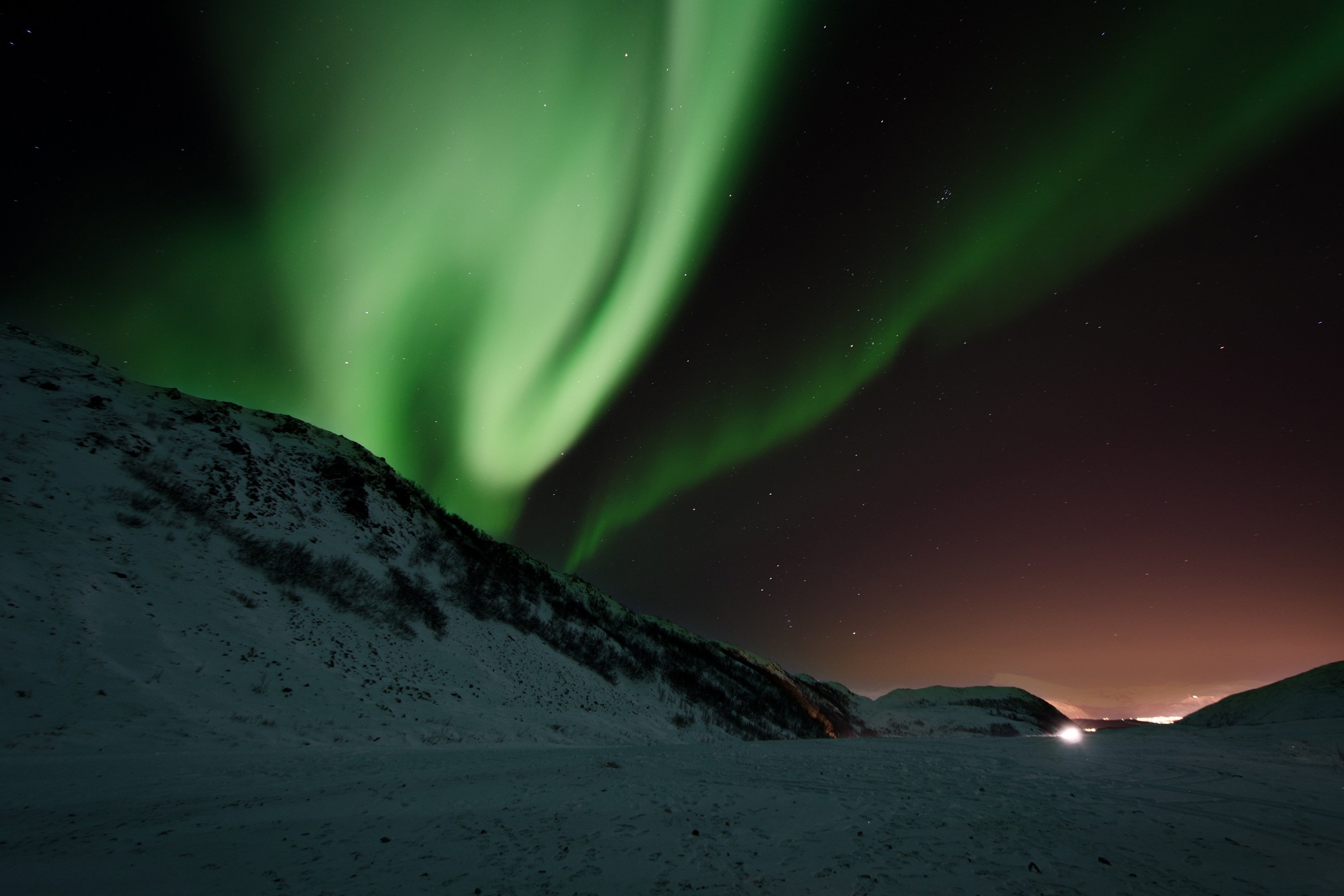 Gr&uuml;ne Nordlichter am Nachthimmel &uuml;ber einer verschneiten Landschaft, am Horizont ein kleines Licht in der Ferne.